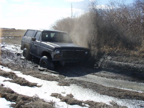 1985 Ford Bronco - A black SUV navigates a muddy trail, kicking up a large spray of mud.