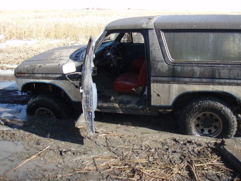 1985 Ford Bronco - This 1985 Ford Bronco is deeply stuck in mud, with its driver's side door ajar, revealing the red interior.