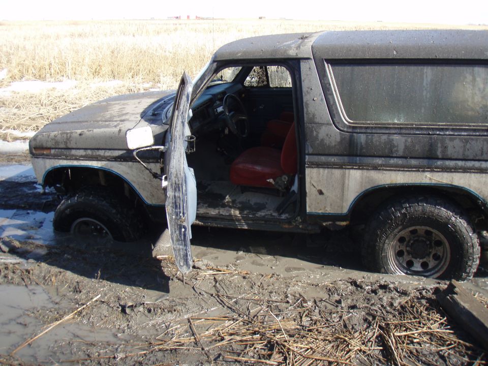 1985 Ford Bronco stuck in mud, driver's door open, red interior visible