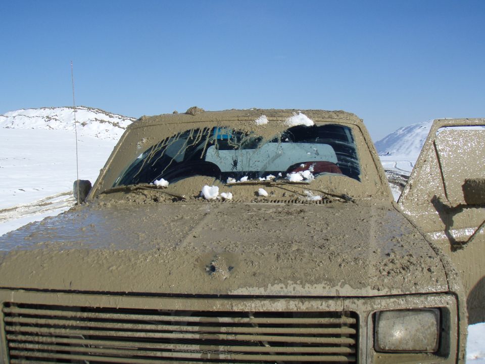 Mud-covered 1985 Ford Bronco front end with snow on windshield. Another vehicle is visible to the right