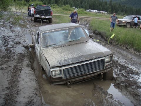1985 Ford Bronco - A muddy pickup truck is stuck in a deep rut, with other vehicles and people in the background on a grassy area.