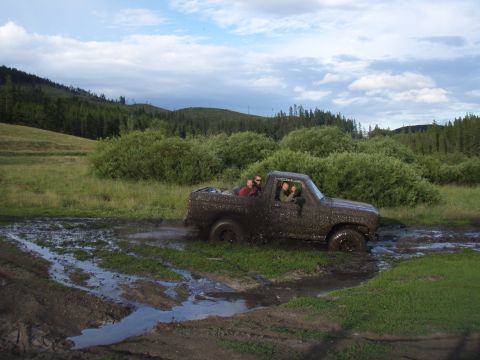 1985 Ford Bronco - This muddy SUV navigates through a challenging off-road terrain, with passengers visible inside the cabin.