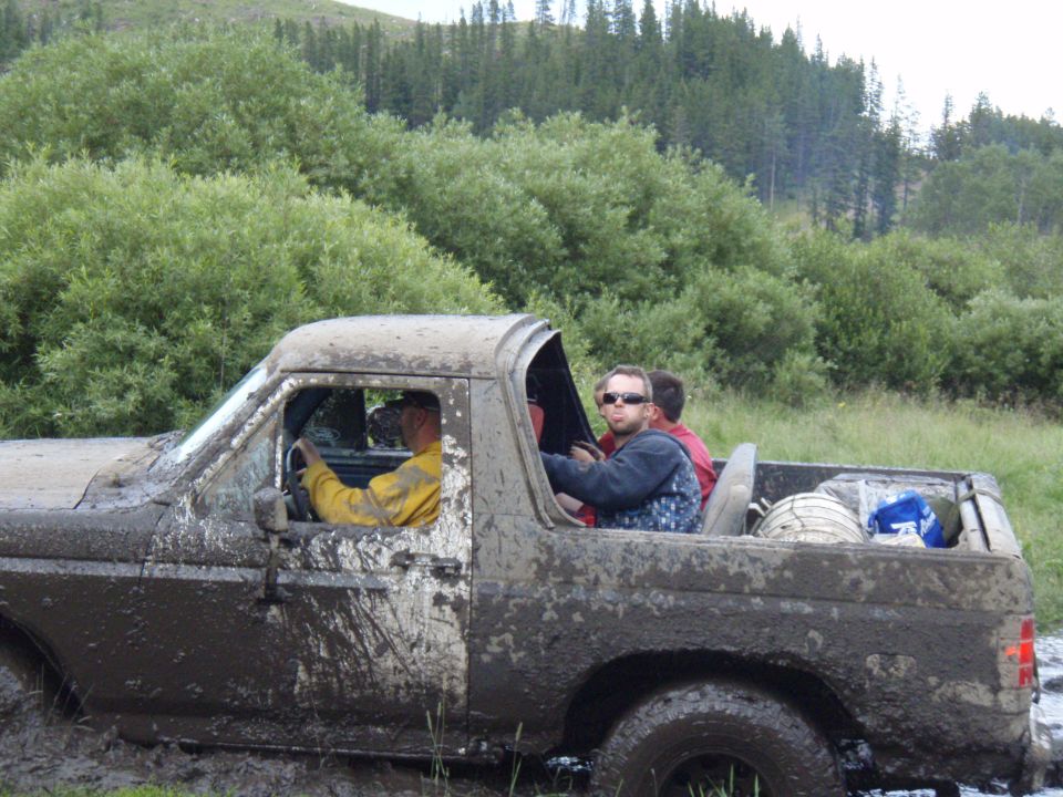 Mud-covered 1980s Ford Bronco stuck in mud with four people inside, driving through a wooded area