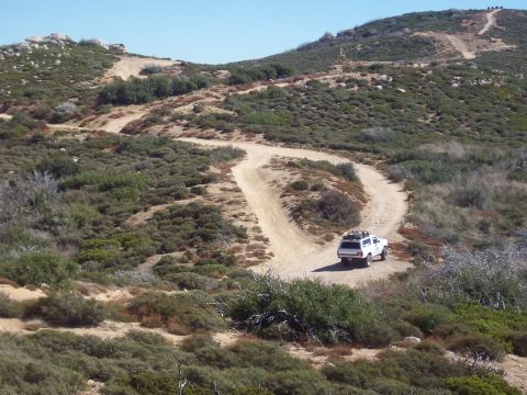 1995 Ford Bronco - A white SUV navigates a winding dirt road through a dry, brush-covered hillside under a clear blue sky.