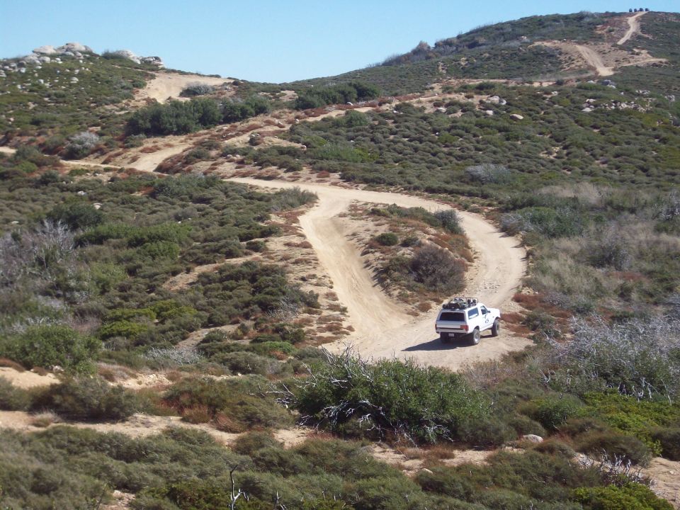 White SUV with roof rack driving on a dirt road in a hilly, scrub-covered landscape