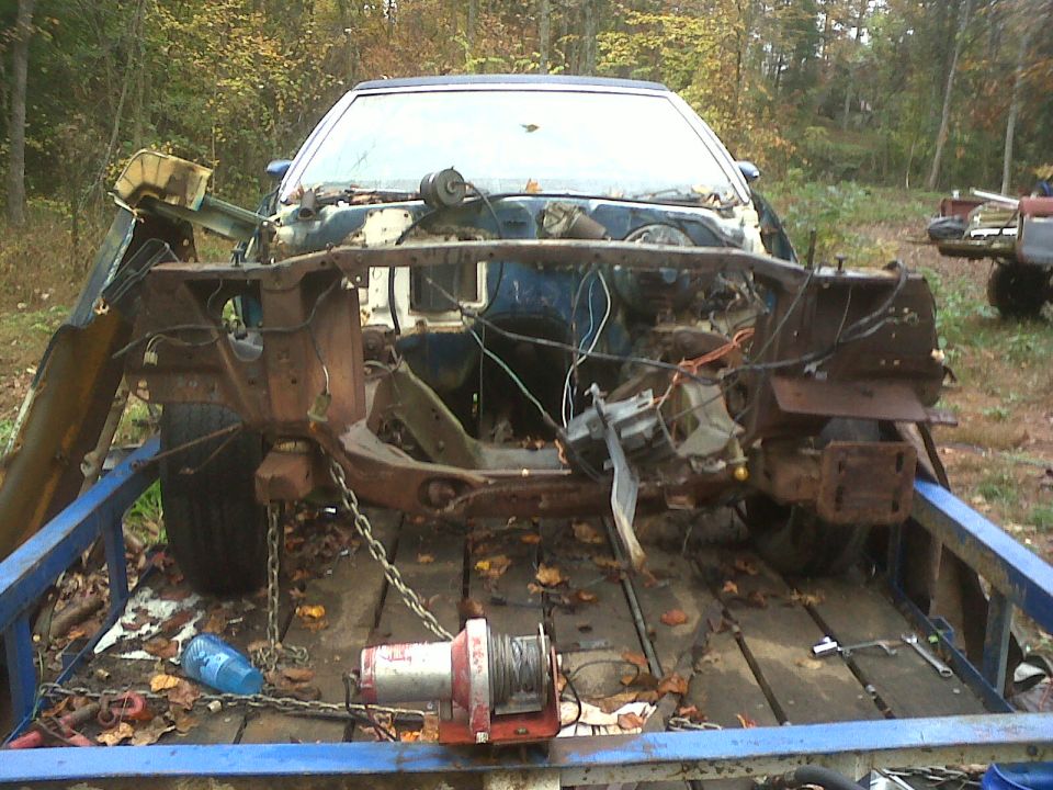Wrecked car chassis on a trailer, front end removed, engine bay exposed, trees in background