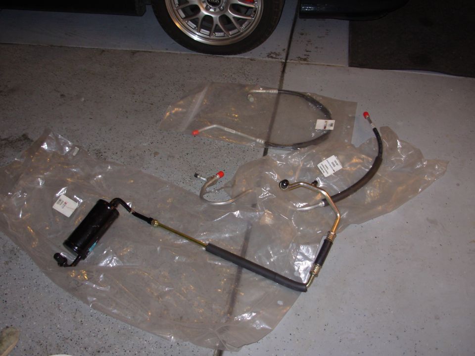 Power steering hoses and reservoir laid out on plastic sheeting, with a car wheel visible in the upper left