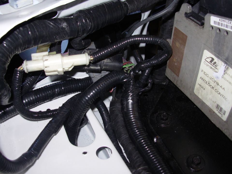 Close-up of a car's engine bay showing a white electrical connector and a bundle of black wires leading to an ABS controller module