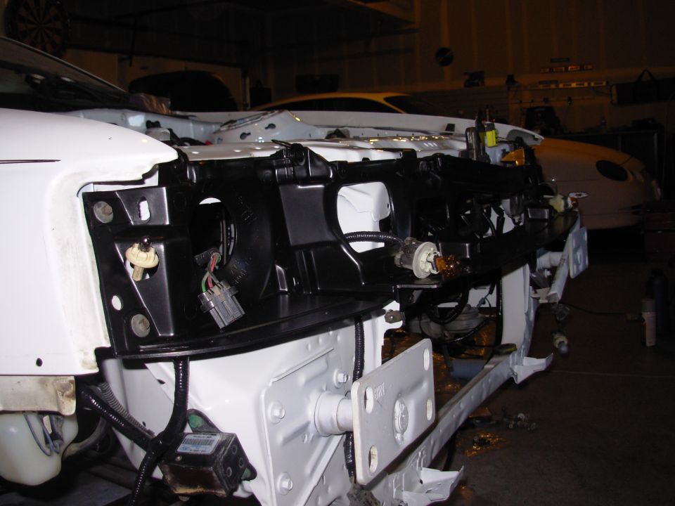 Car front end disassembled, showing wiring and internal components. Another white car is visible in the background