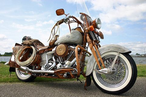 1973 Ford Ranchero - A custom motorcycle with extensive leather detailing and a western theme is parked on a paved surface near a grassy bank and water.