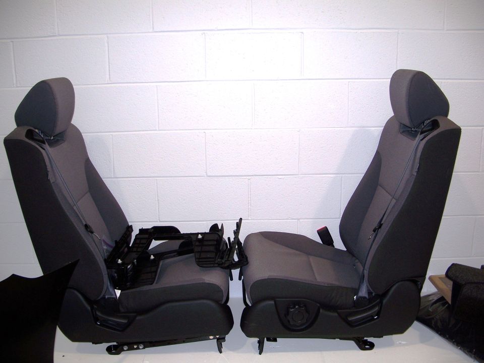 Two gray car seats with black trim and seatbelts against a white brick wall