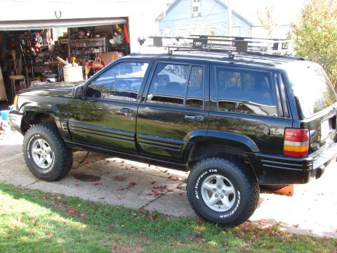 1996 Jeep Grand Cherokee - This black 1996 Jeep Grand Cherokee is parked in front of an open garage, equipped with a roof rack and off-road tires.