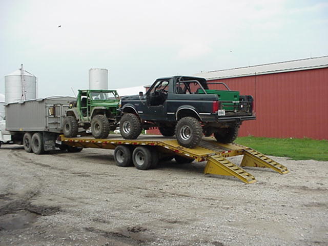 1992 Ford Bronco and a green Jeep on a flatbed trailer, parked on gravel near a red barn