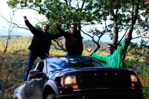 1996 Ford Bronco - Three friends celebrate on top of a black Ford F-150 Raptor in a scenic, wooded landscape.
