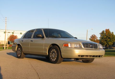 2004 Ford Crown Victoria - A gold sedan is parked on a road with trees and buildings in the background.