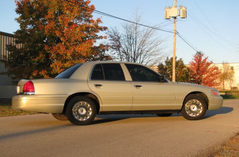 2004 Ford Crown Victoria - A gold sedan is seen from the side on a street with trees in the background.
