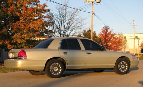 2004 Ford Crown Victoria - Side profile of a tan 2004 Ford Crown Victoria sedan parked on a paved surface with trees in the background.