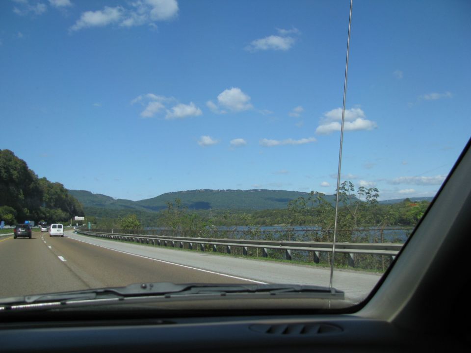 View through car windshield of highway along a river with wooded ridges