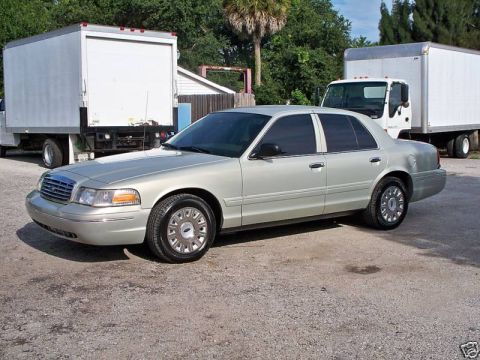 2004 Ford Crown Victoria - Silver sedan parked on gravel lot with two white box trucks behind it.