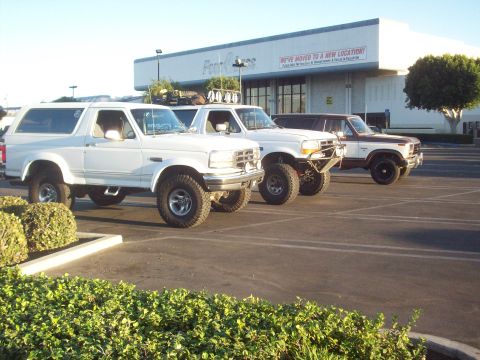 1995 Ford Bronco - Three Ford Broncos are parked in a lot. The building behind them has a sign indicating a new location.