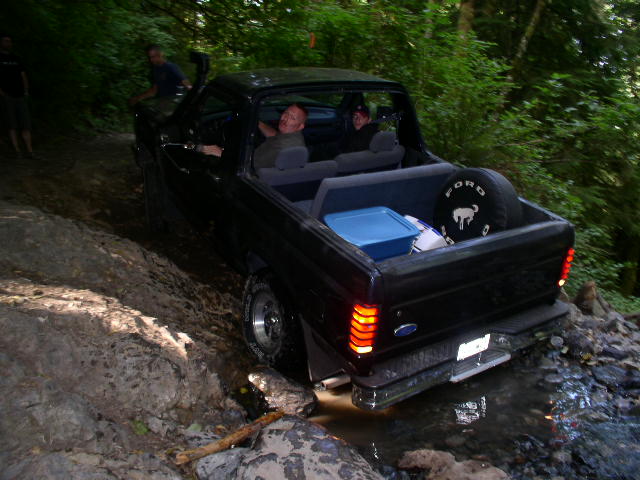 Topless dark blue Ford Bronco in a creek with Ford spare tire cover and cooler in back