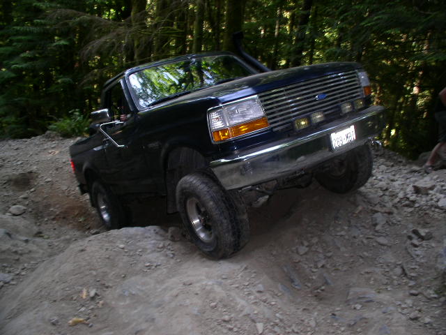 Dark blue doorless Ford Bronco flexing over a dirt mound on a forest trail