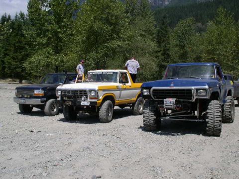 1993 Ford Bronco - Three off-road vehicles are parked on a gravel lot with trees and mountains in the background.