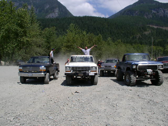 Row of lifted Ford 4x4 trucks and a Bronco on gravel at an off-road meet