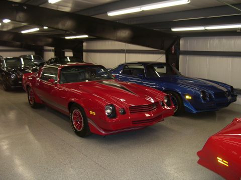 1979 Chevrolet Camaro - Two Chevrolet Camaros, one red and one blue, are parked in a garage with overhead lighting.