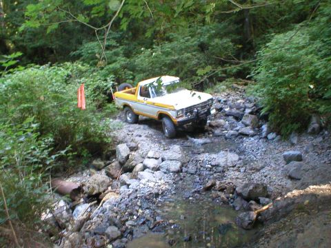 1978 Ford Bronco - 1978 Ford Bronco driving through a rocky stream, surrounded by dense foliage.