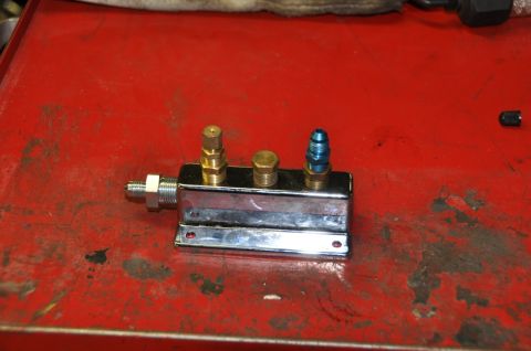 1996 Ford Bronco - A chrome manifold with three fittings, including a brass fitting, a capped fitting, and a blue anodized fitting, rests on a red surface.