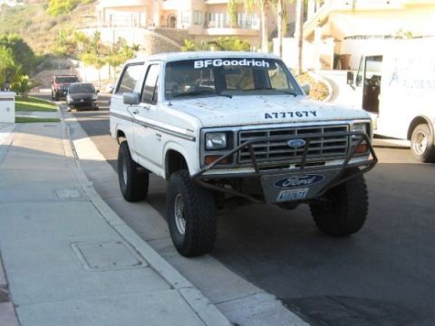 1986 Ford Bronco - White full-size Ford Bronco parked at a suburban curb, wearing a tube prerunner front bumper with Ford logo, a BFGoodrich windshield banner…