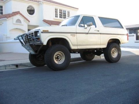 1986 Ford Bronco - A white first-generation-style full-size Ford Bronco SUV shown from a front three-quarter angle, lifted on chrome-style wheels with large o…