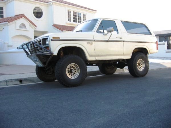 White lifted Ford Bronco SUV with brush guard, parked on a street in front of a stucco house.