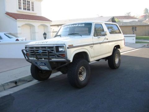 1986 Ford Bronco - A white 1980s full-size Ford Bronco with a black tube front bumper and Ford-lettered skid plate sits in a suburban driveway, front three-qu…
