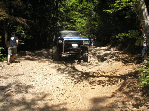1978 Ford Bronco - A blue off-road SUV navigates a challenging rocky path through a wooded area.