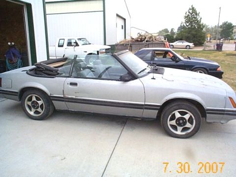 1984 Ford Mustang - A silver convertible Mustang is parked outside a garage with other vehicles nearby.