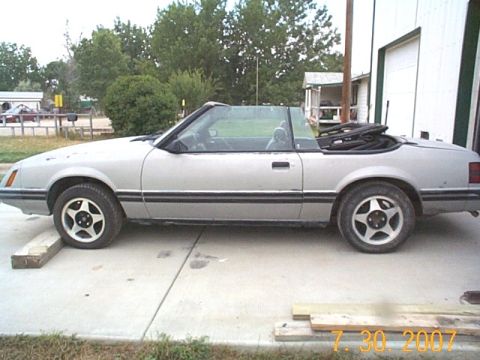 1984 Ford Mustang - A silver convertible is parked on a concrete surface with a garage in the background.