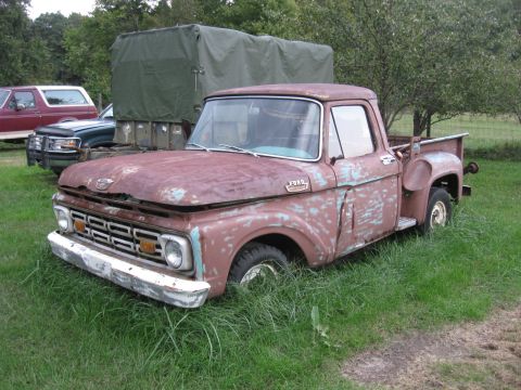 1966 Ford F-250 - A weathered Ford F-series truck sits in overgrown grass next to other vehicles and a large tarp-covered structure.