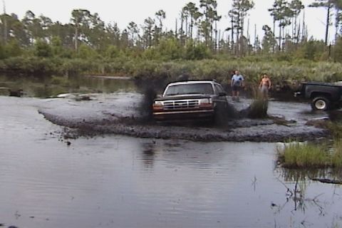 1993 Ford Bronco - A 1993 Ford Bronco navigates a muddy water obstacle with another truck visible in the background.