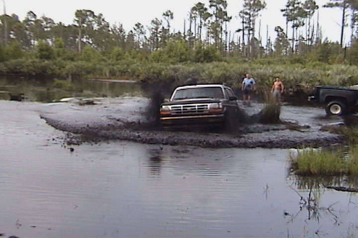 White Ford Bronco driving through swamp mud, spraying water, with onlookers and a pickup on the bank