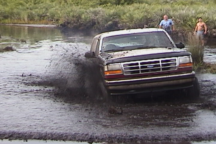 Two-tone Ford Bronco splashing through a muddy water hole with spectators watching