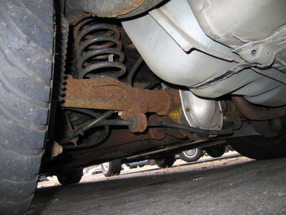 Underside view of a vehicle's rear axle, coil spring, and tire. Other vehicles are visible in the background