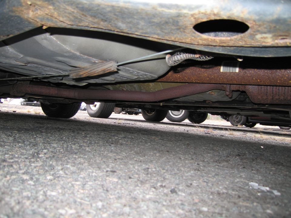 Underside of a car showing rusty frame, exhaust pipe, and wheels of multiple vehicles parked behind