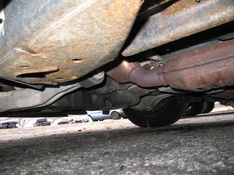 1991 Ford LTD Crown Victoria - The underside of a vehicle reveals its exhaust system and heavily rusted frame components, with another car partially visible in the distan…