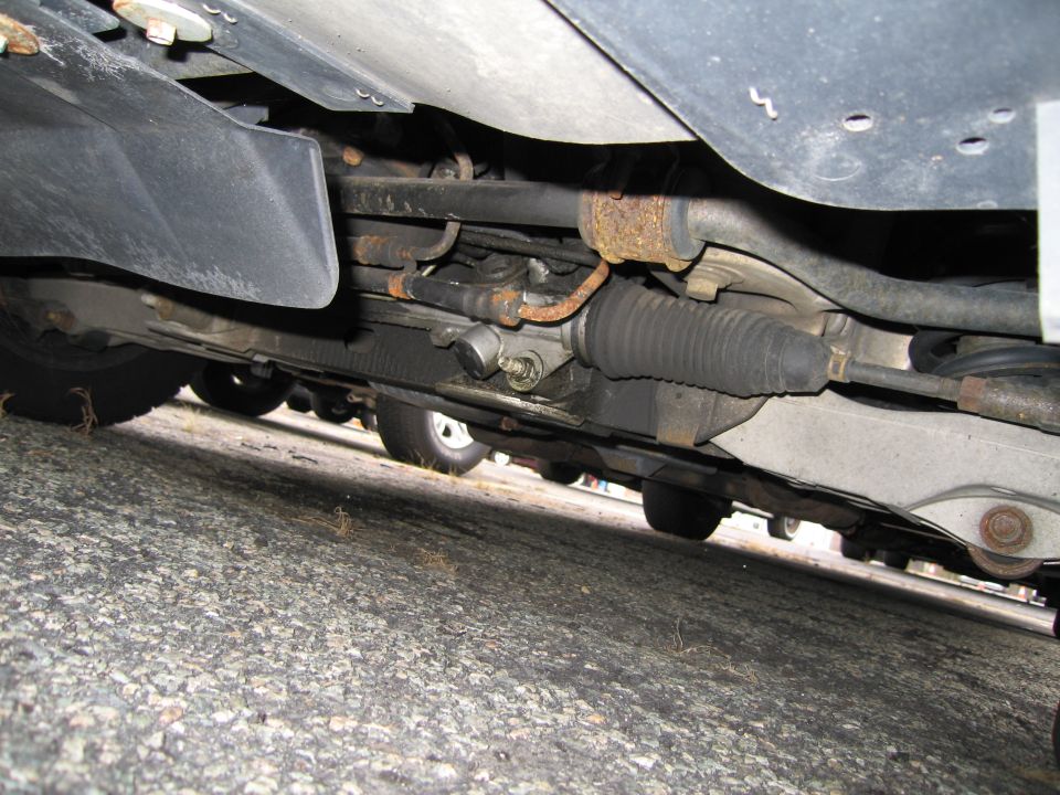 Underside of a car showing the steering rack and suspension components. Other vehicles are visible in the background