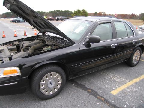 1991 Ford LTD Crown Victoria - Black sedan with hood open, parked in an asphalt lot with traffic cones and other vehicles visible.