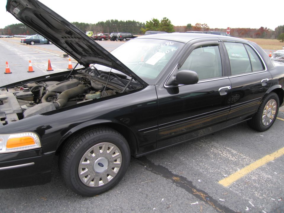 Black sedan with hood open, showing engine. Visible text on windshield. Police car and other vehicles in background