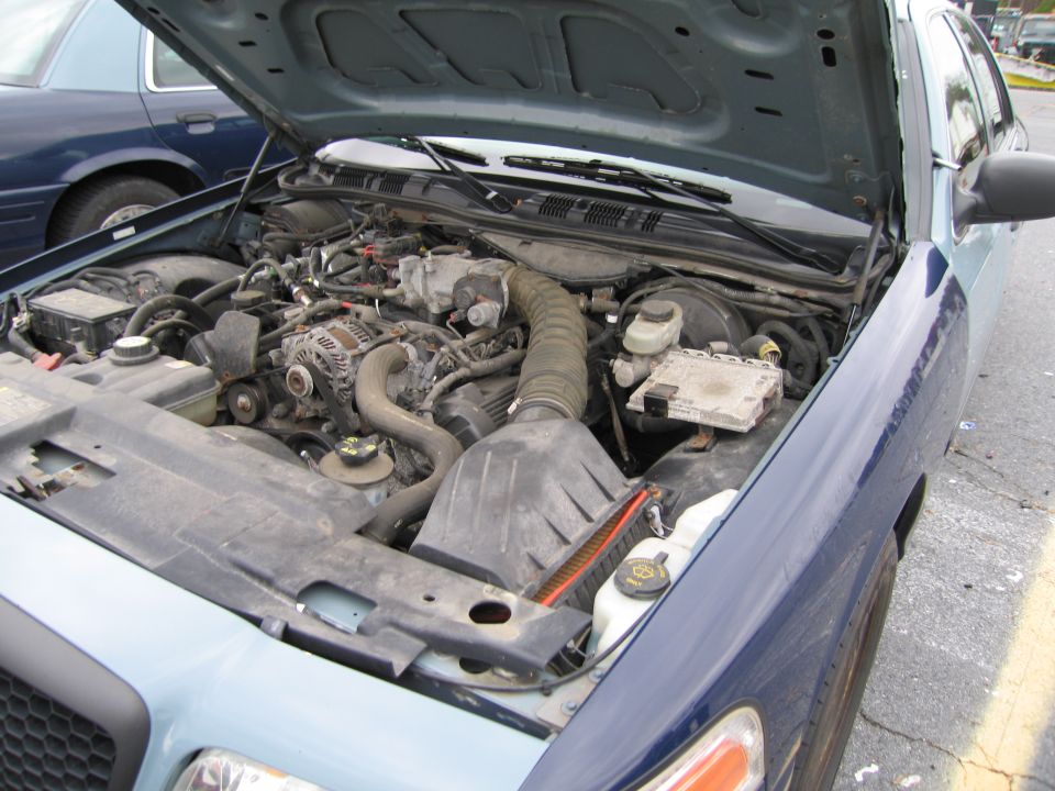Engine bay of a blue Ford Crown Victoria, hood open. Another blue Ford Crown Victoria is visible to the left