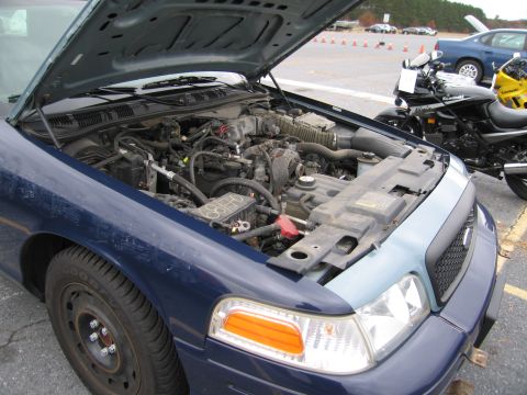 1991 Ford LTD Crown Victoria - The engine bay of a blue Ford Crown Victoria is visible, with a motorcycle parked to the right.